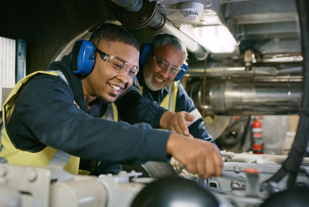 Two smiling men, in uniform and working on engine repair in factory wearing safety glasses. Senior expert mechanic coaching a young guy in a manufacturing plant. Professional mechanic in development